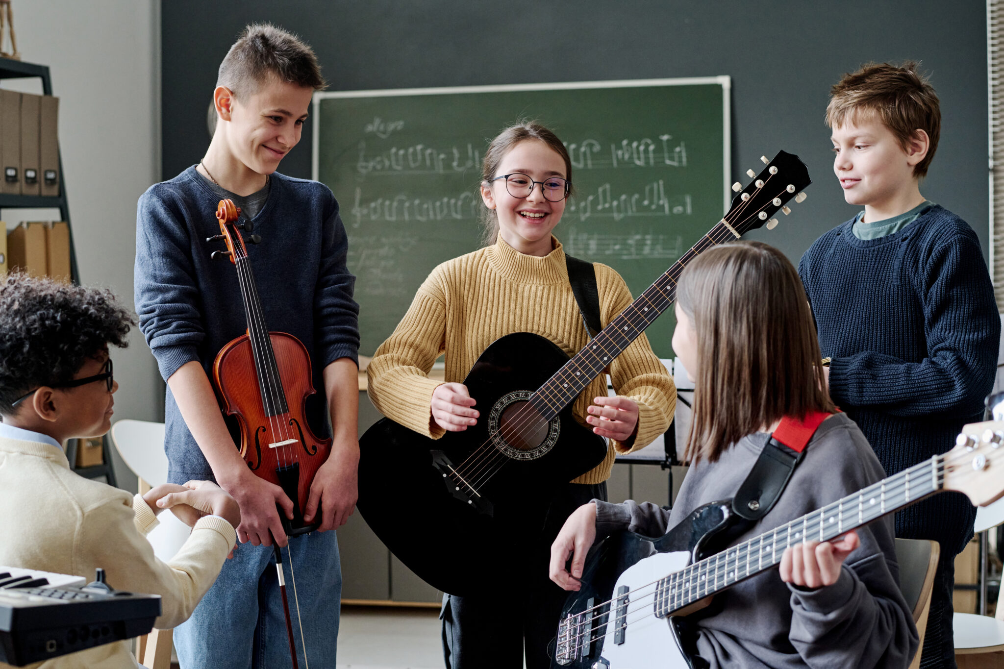 Eine Gruppe von jüngeren Schülern unterhält sich im Klassenraum, jeder von ihnen hält ein anderes Instrument in der Hand, wie Keyboard, Geige und verschiedene Gitarren